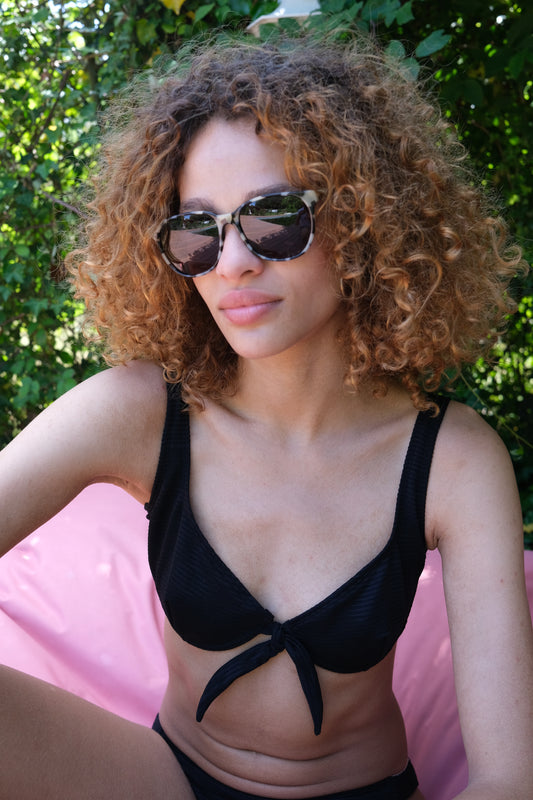 Woman sitting on pink beanbag in front of bushes in a PRISM bikini